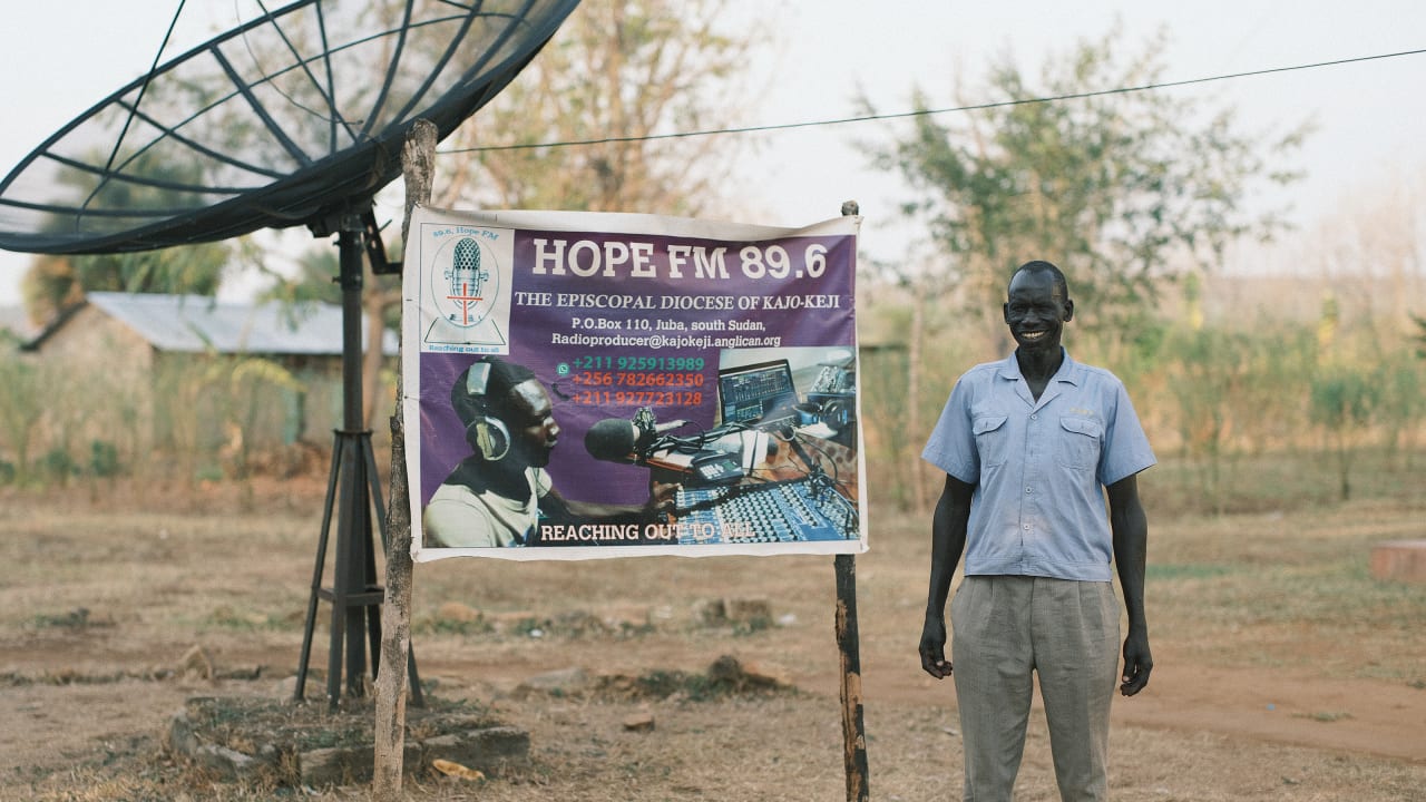 Man stands next to Hope FM 89.6 banner with a satellite dish and house in the background.