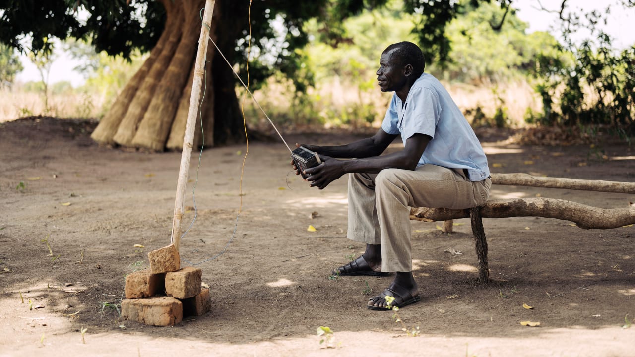 Man sits on a bench outside, listening to a radio with makeshift antenna.