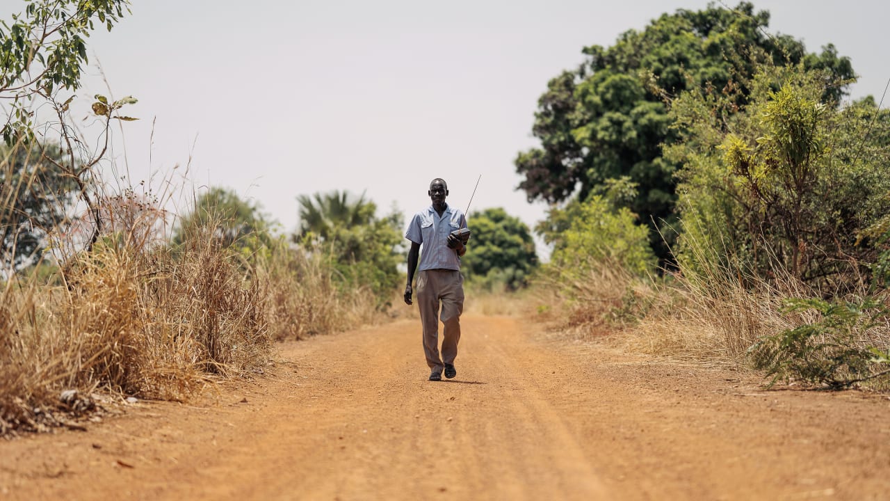 Man walks down dirt road with radio in South Sudan, surrounded by grass and trees.