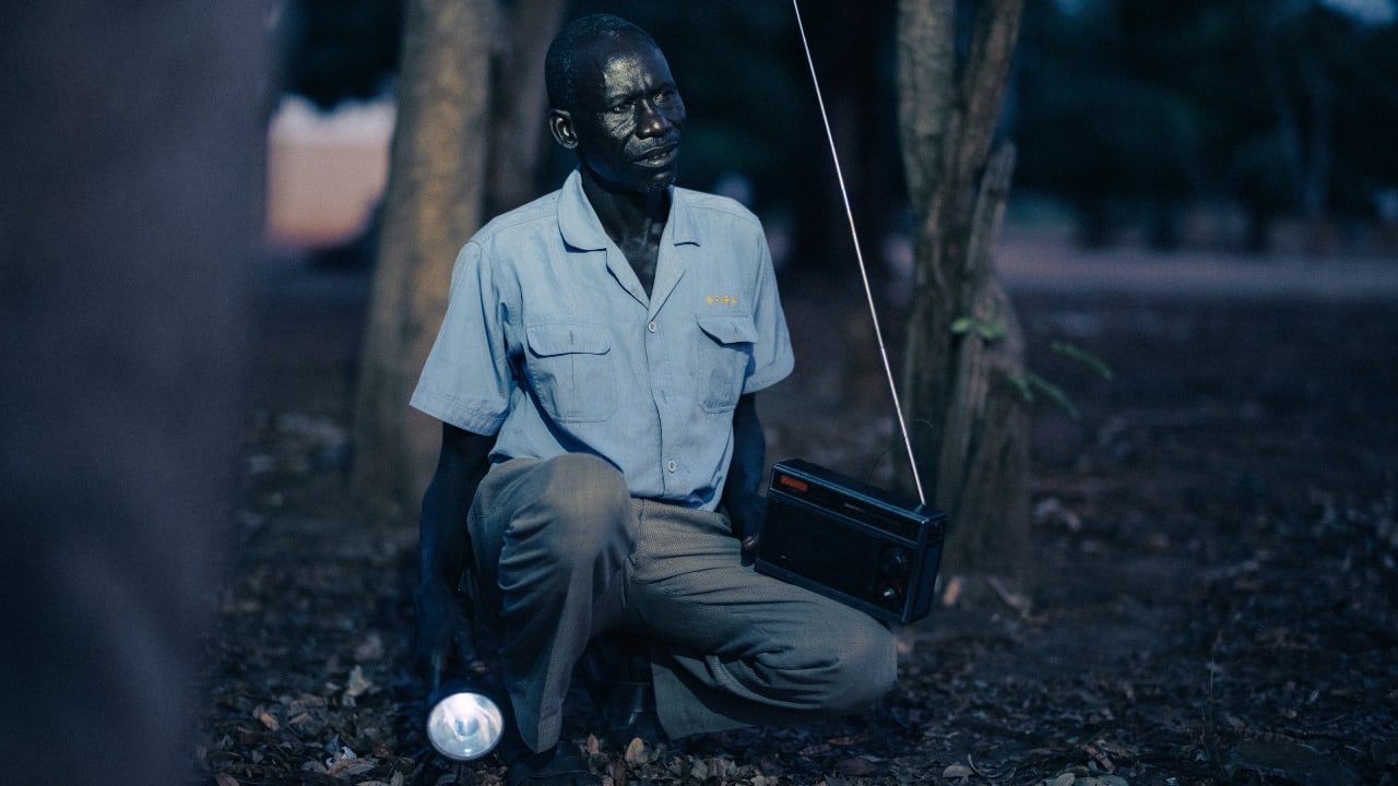 Man with a radio and flashlight squats between trees, looking towards the horizon.