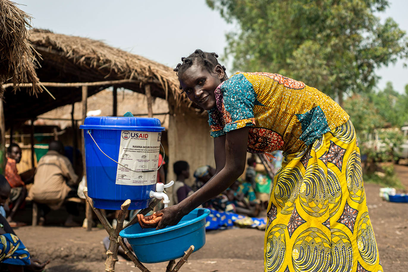 A woman in the DRC washes her hands using a bowl and water bucket on a stand.