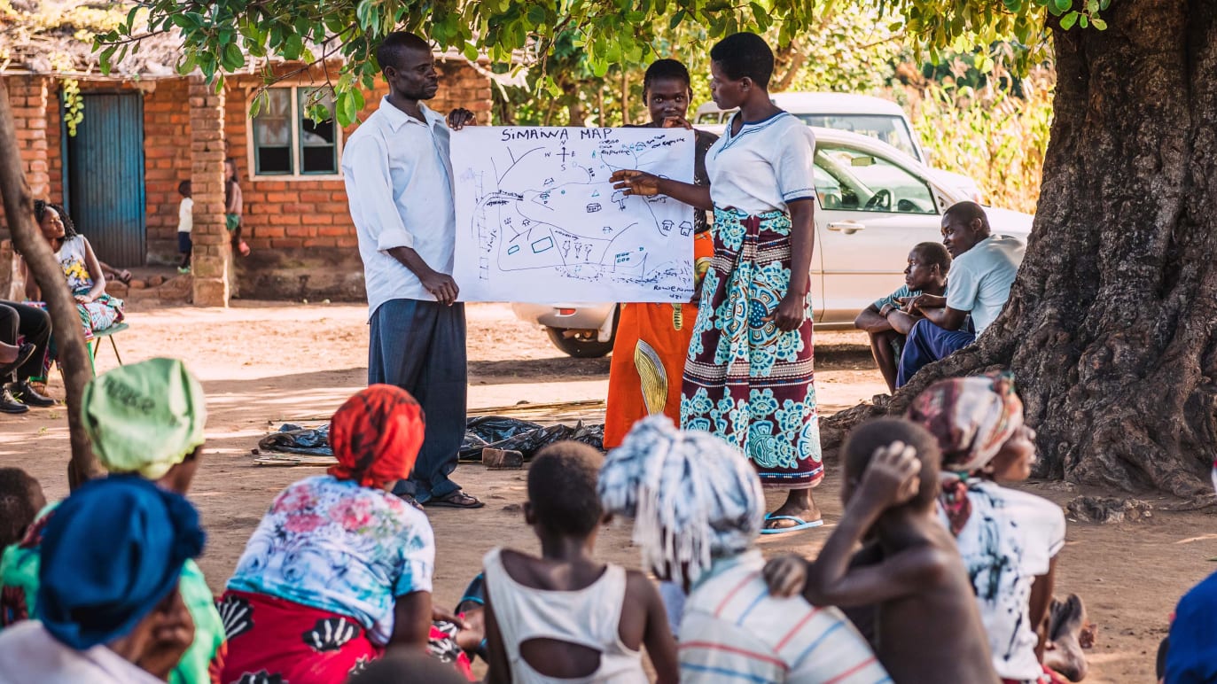 Des membres d’une communauté assis par terre, en plein air, regardent une facilitatrice qui leur montre quelque chose sur un tableau