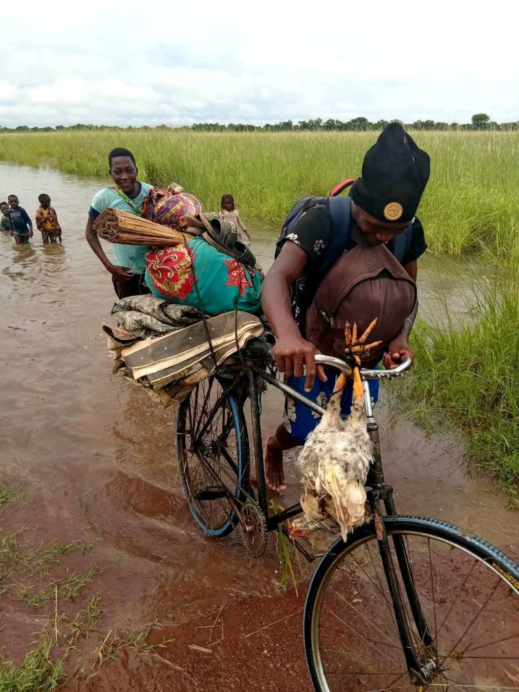 Man pushes overloaded bike through flood waters with chicken hanging on handle bars.