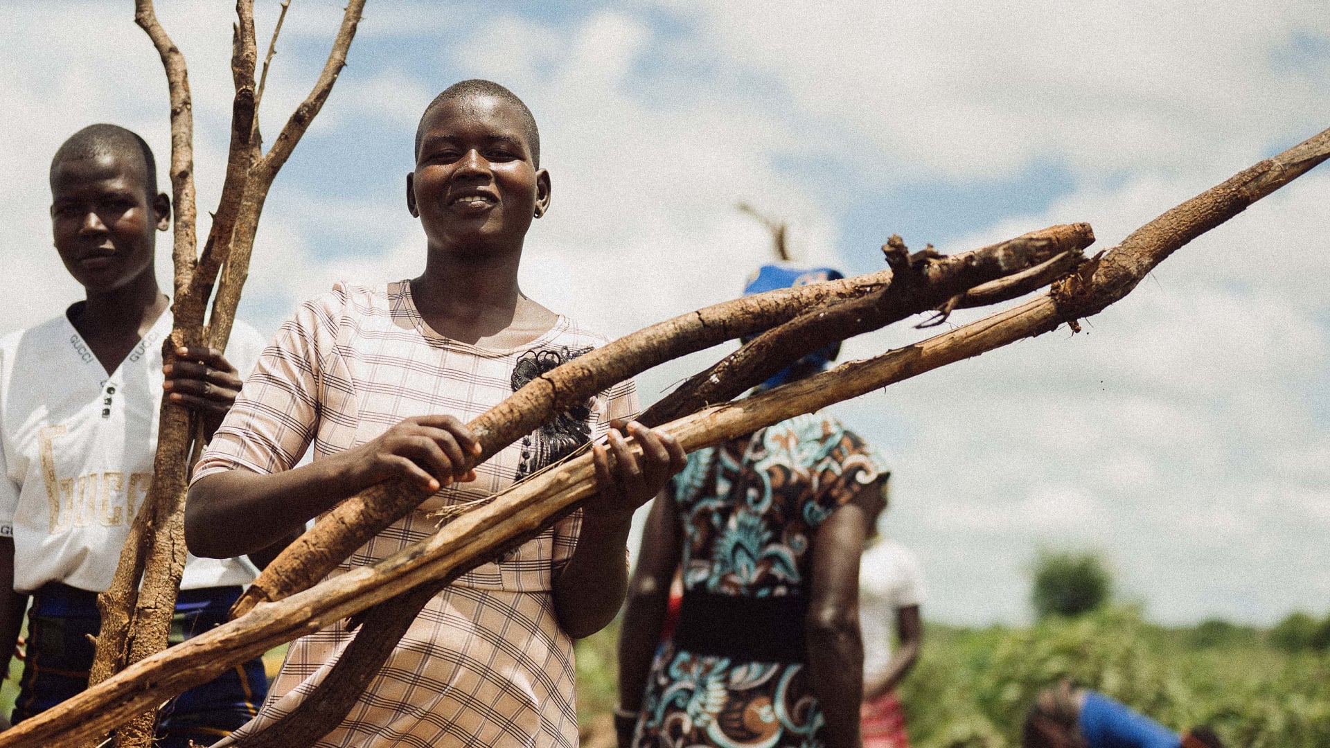 A young woman with a shaved head smiles, holding several thick branches, alongside a second young person holding a branched stick. Other blurred figures and green plants are in the background under a cloudy sky.