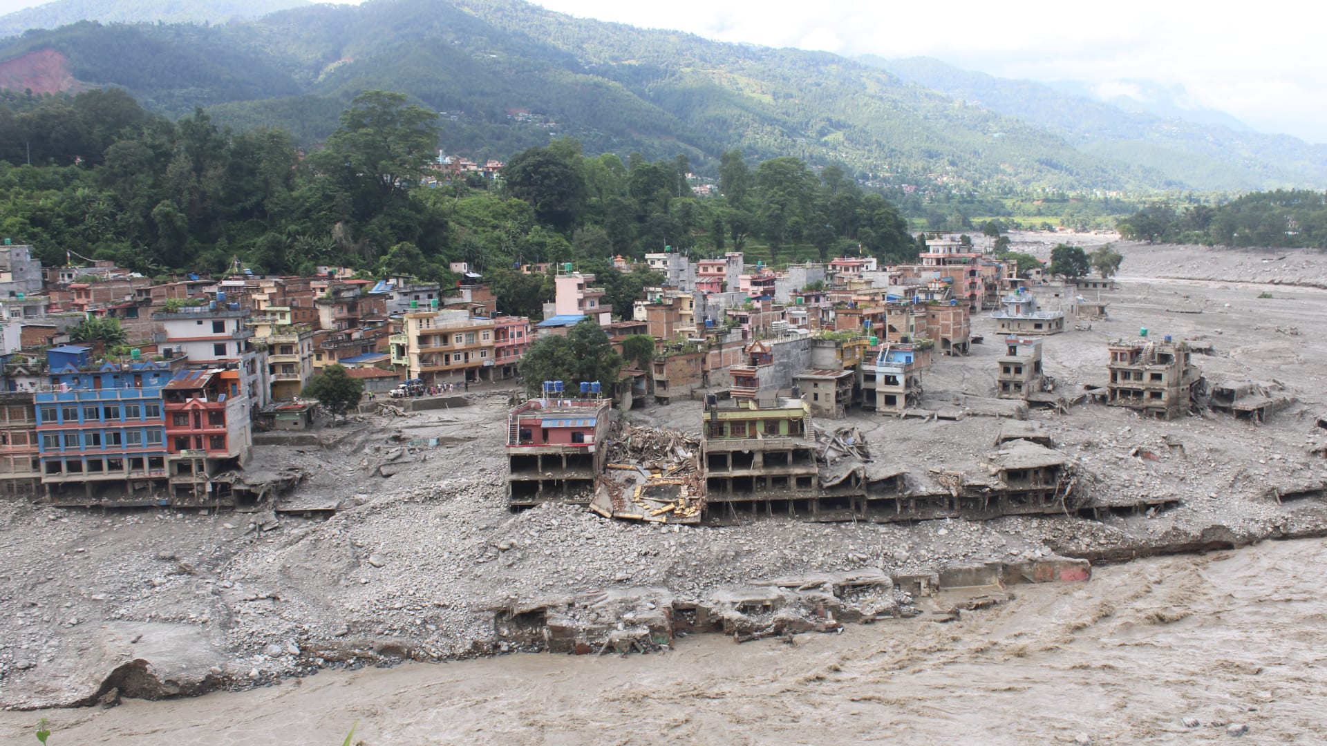 Many multi-storey buildings in Nepal are partially buried in grey mud and debris, with several collapsed structures, alongside a wide muddy river. Green hills and mountains rise in the background.
