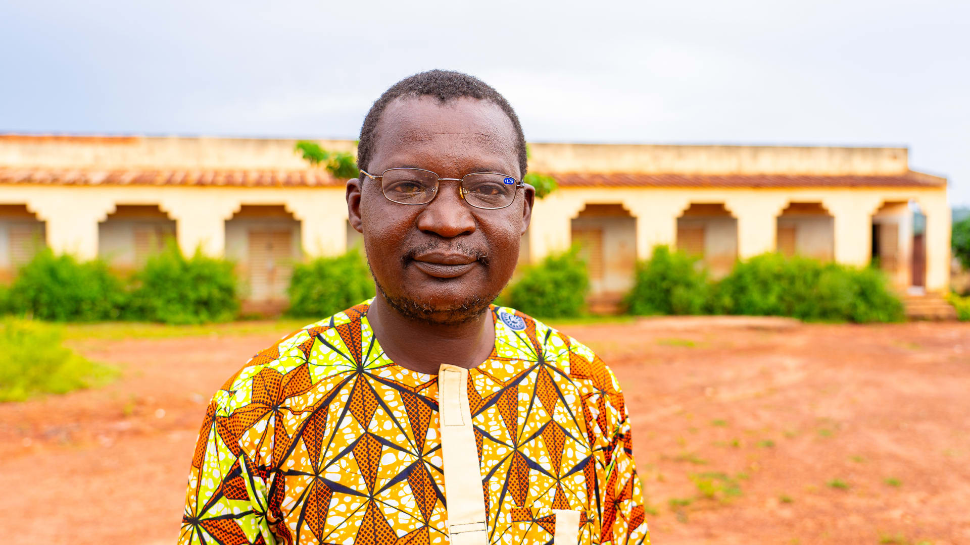 Portrait of a man named Mahamadou wearing glasses and a colorful shirt, standing outdoors in front of a building.