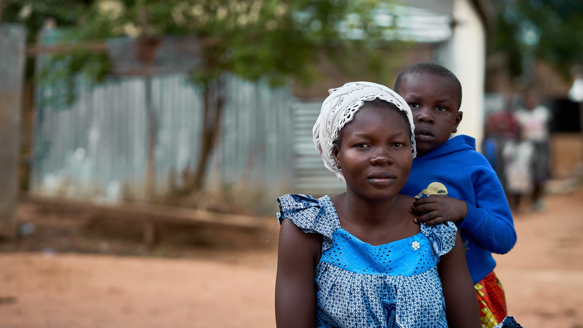 Julia in colourful patterned clothing sits looking into the camera outside in her village, while her son stands leaning against her.