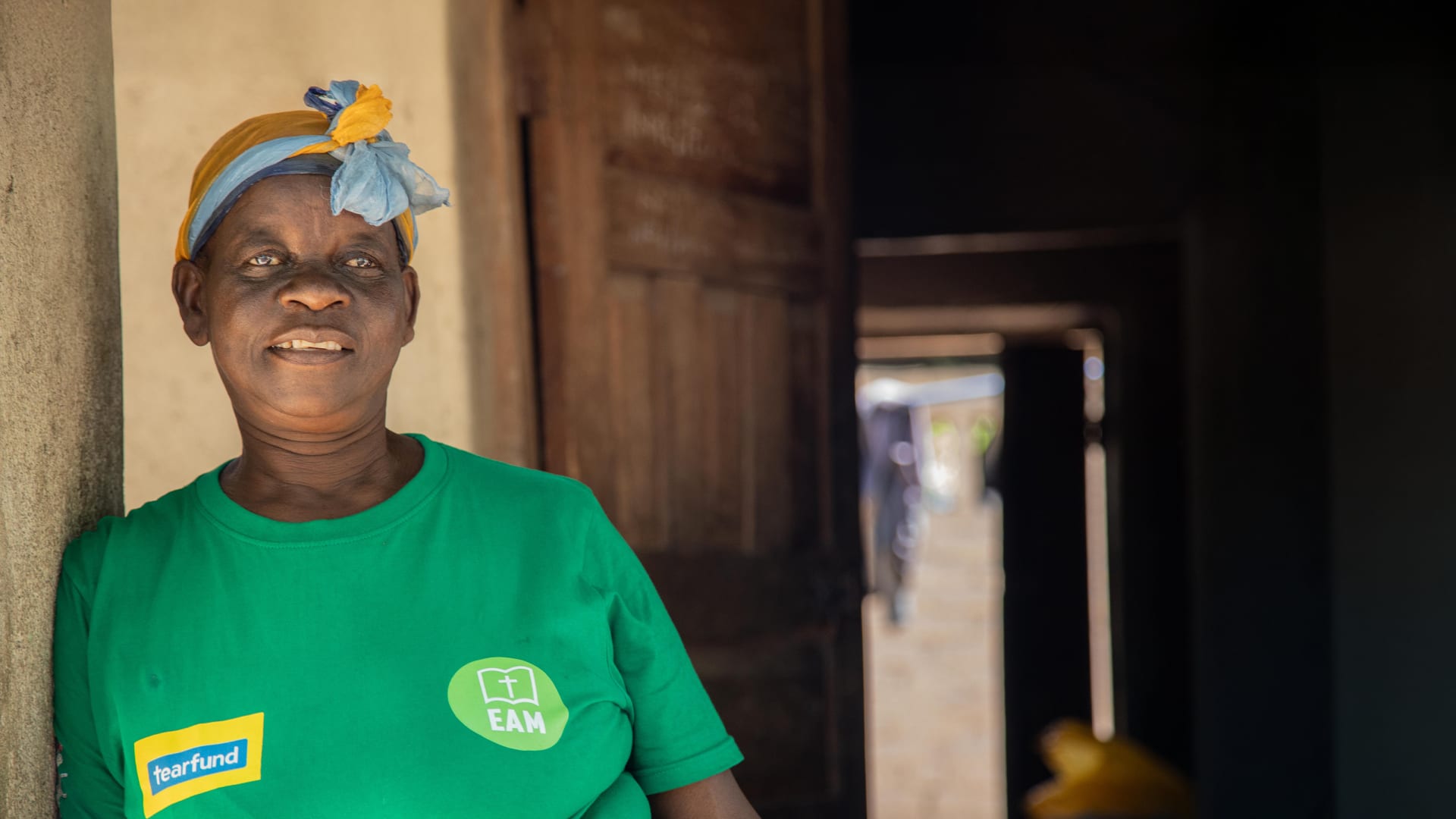 Woman in green Tearfund shirt and headscarf smiles in doorway. Building in background.