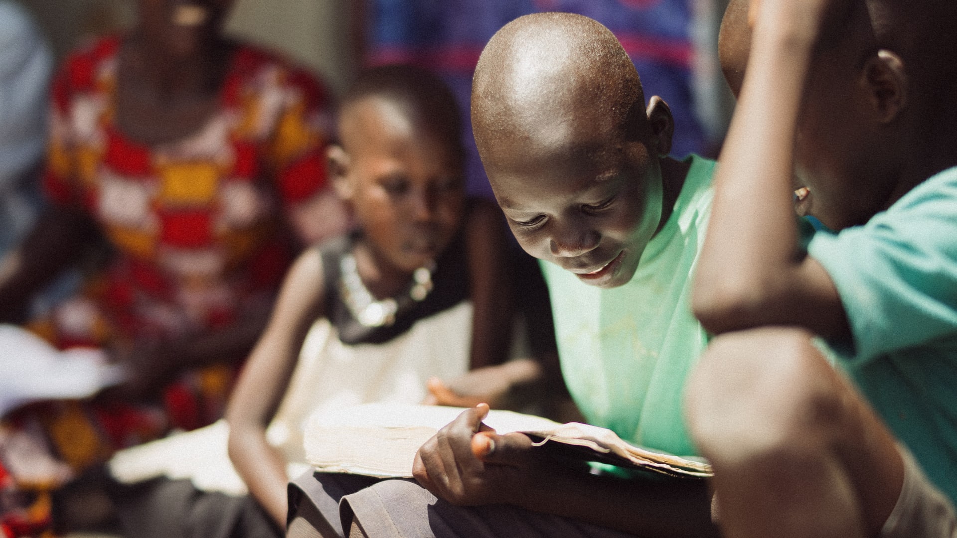 Young African child engrossed in reading a book, with other children nearby.