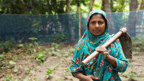 A woman wearing a teal headscarf and floral tunic looks at the camera, holding a hoe over her left shoulder. She stands in a field with loose soil and green plants.