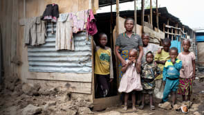 A woman (Rose) and her children (seven) stand in front of a wooden structure. Clothes hang some corrugated metal on the side of the building.