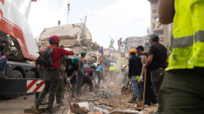 Rescuers and volunteers search through rubble from a collapsed building, crane nearby.