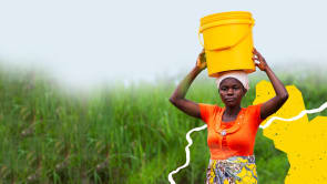 A women wearing orange is carrying a yellow bucket on her head.