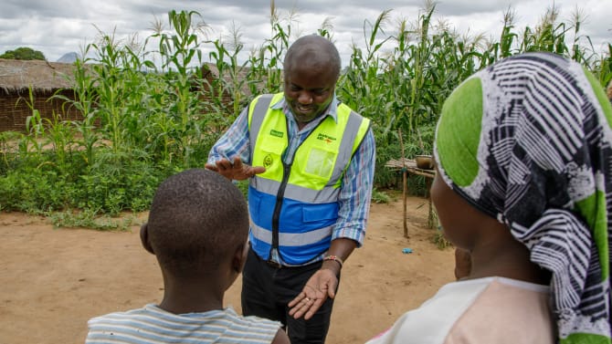 A man in a yellow and blue reflective vest speaks to a child and an adult in a rural village. He gestures with an open palm holding a small object, against a background of maize plants and thatched huts.