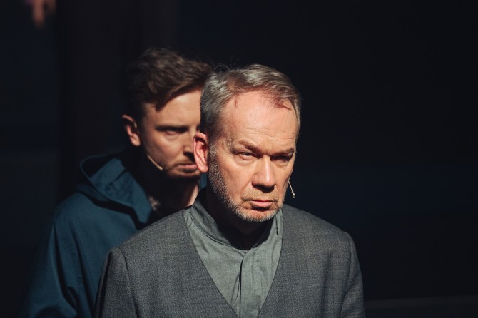 Zbliżenie na dwóch mężczyzn - starszy (Mirosław Baka) stoi ze zmarszczonymi brwiami. Za jego plecami młodszy mężczyzna (Piotr Biedroń) z podobnie chmurną miną. Dookoła mrok.