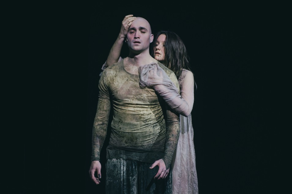 Na zdjęciu dwie osoby (Marek Puchowski, Maria Wróbel) na scenie teatralnej. Jedna stoi z zamkniętymi oczami, druga obejmuje ją od tyłu, dotykając głowy. Oświetlenie punktowe, ciemne tło.