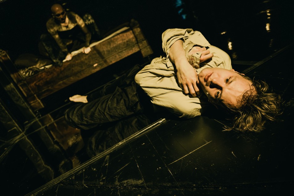 Na zdjęciu osoba (Robert Ciszewski) leżąca na błyszczącej podłodze sceny, zwrócona twarzą do obiektywu. W tle widoczna druga osoba (Marek Puchowski) pochylona nad obiektem.