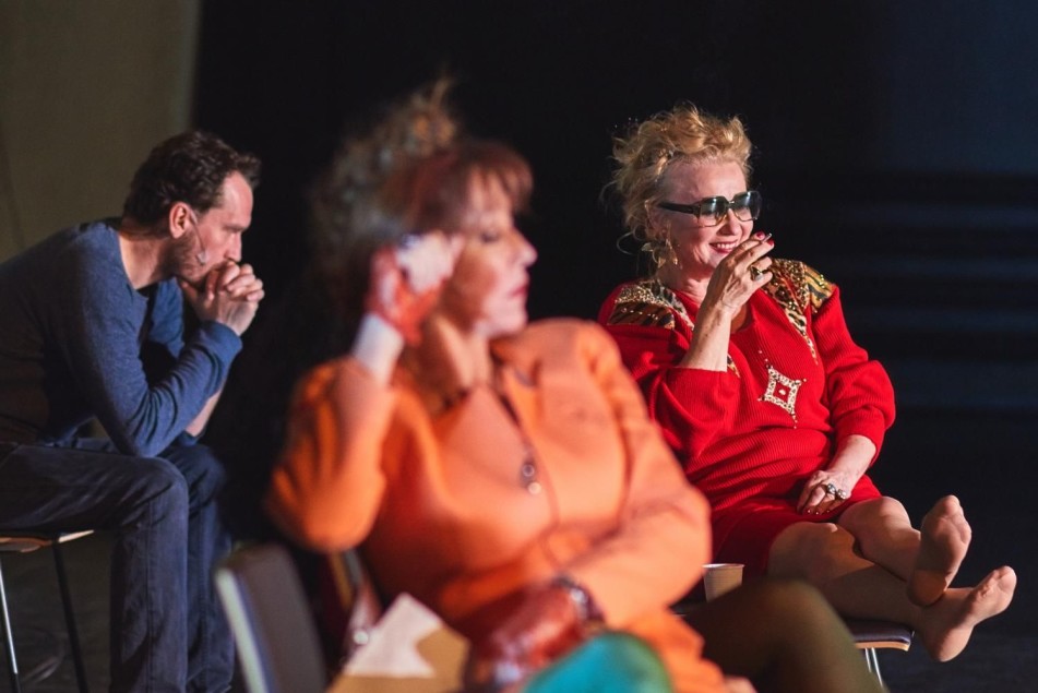 Trzy osoby siedzą na krzesłach na scenie. Na drugim planie kobieta w czerwonej sukni i okularach pali papierosa, uśmiechnięta. Przed nią kobieta w pomarańczowym stroju, zamyślona. W tle mężczyzna siedzi pochylony, z rękami splecionymi przy twarzy.