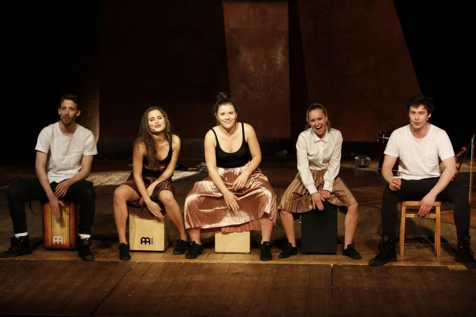 Pięcioro aktorów siedzi na drewnianej scenie na skrzynkach i stołku, zwróceni do widowni. Ubrani w jasne, sceniczne kostiumy, niektórzy uśmiechnięci. W tle prosta, geometryczna scenografia w ciepłych brązach.