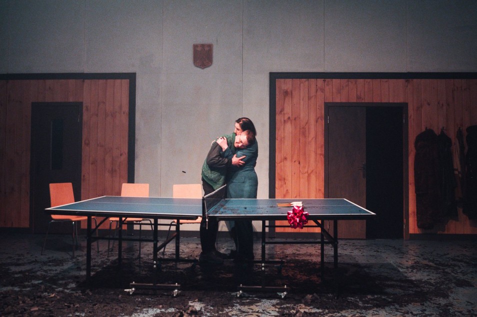 Dwoje aktorów przy stole do ping-ponga obejmuje się na środku sceny w surowej scenografii.