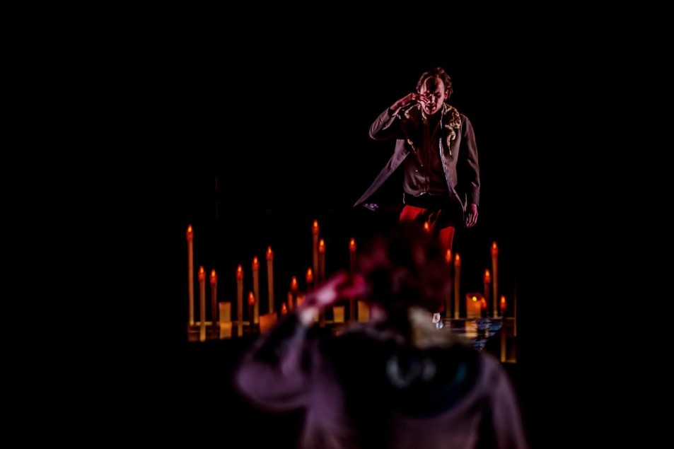 Aktor stoi na podwyższeniu w ciemnej przestrzeni scenicznej, oświetlony punktowym światłem. W tle i wokół niego widoczne są zapalone świece, tworzące rytmiczny, symboliczny układ. Na pierwszym planie rozmyta sylwetka innej postaci.