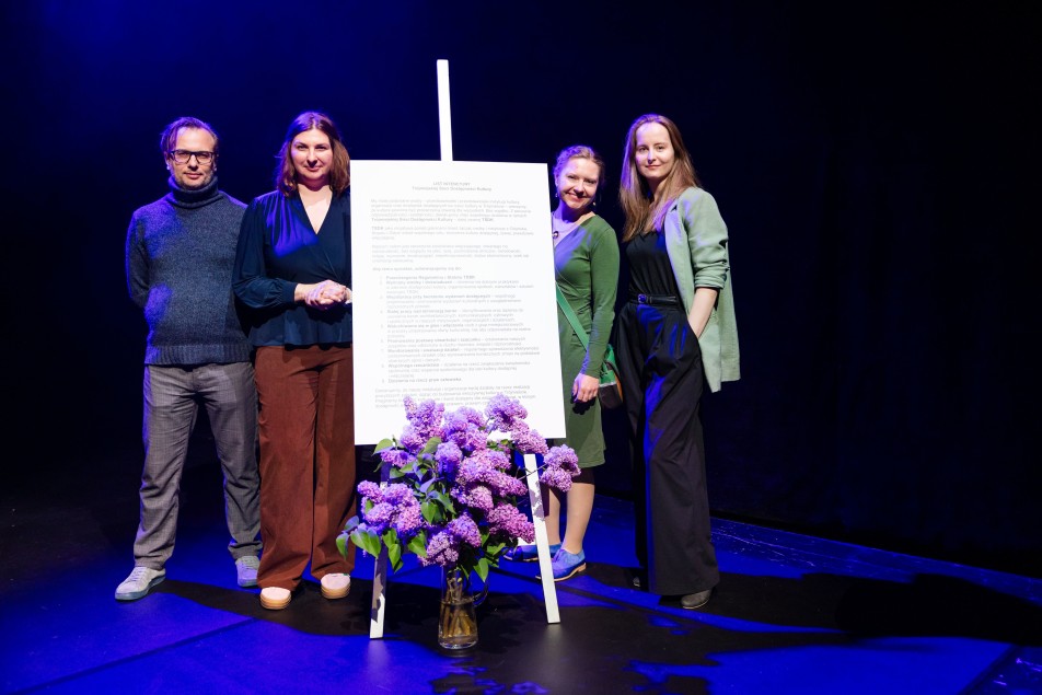 Cztery osoby stoją na scenie w niebieskim świetle reflektorów obok dużej tablicy z tekstem, to pracownicy Teatru Wybrzeże. Dwie kobiety w środku uśmiechają się i wskazują na planszę, na której znajduje się tekst listu intencyjnego. 