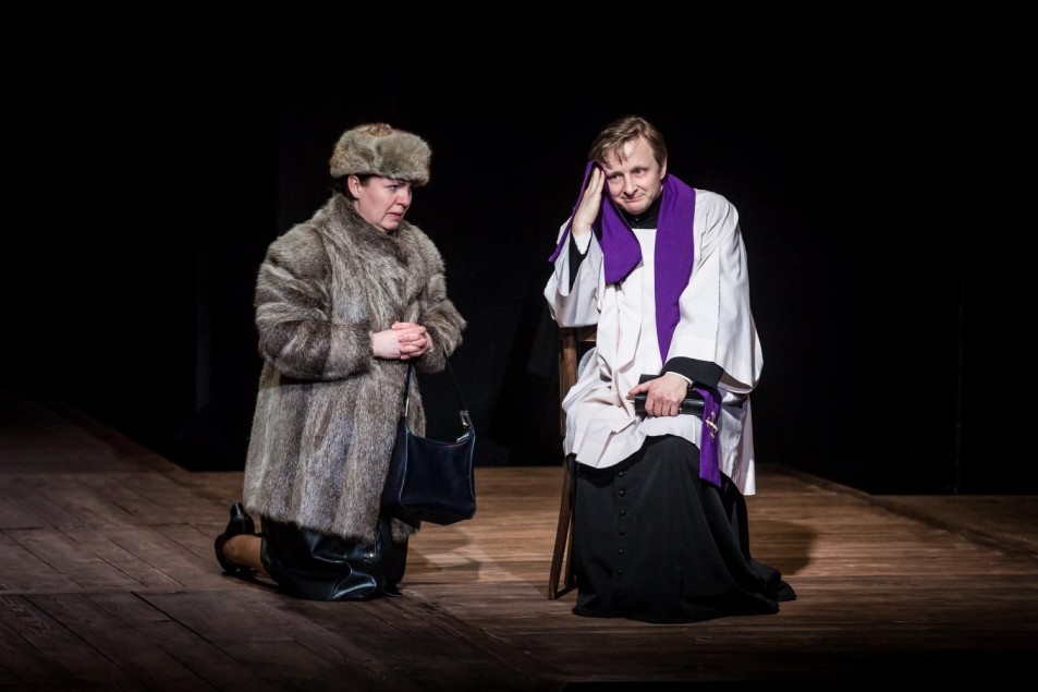 Scena teatralna: kobieta w futrzanym płaszczu klęczy obok siedzącego księdza w białej szacie z fioletową stułą. Postacie skupione, atmosfera napięcia i intymnej rozmowy.