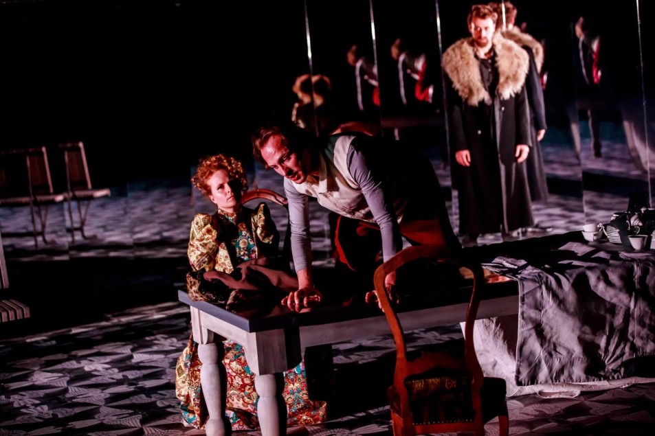 Scena teatralna z kilkoma postaciami. Na pierwszym planie mężczyzna pochyla się nad stołem, obok siedzi kobieta w bogato zdobionej sukni.