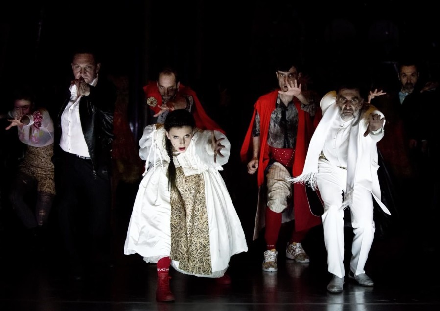 Grupa aktorów na ciemnej scenie wyciąga dłonie w stronę widza; centralnie kobieta w jasnym kostiumie i czerwonych butach.