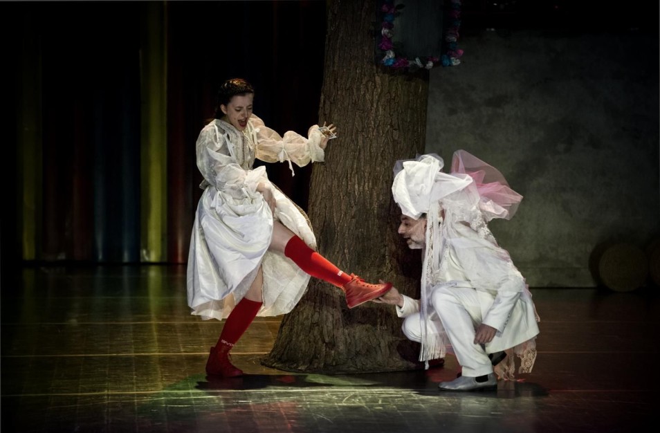 Kobieta w białej sukni opiera stopę o pień drzewa, obok klęczy mężczyzna w białym kostiumie z welonem.