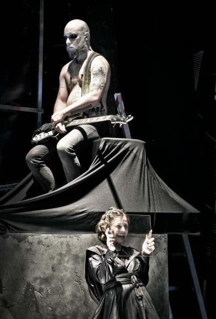 Mężczyzna z gitarą siedzi na podwyższeniu, poniżej kobieta trzyma przezroczystą taflę szkła przed twarzą.