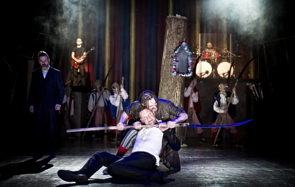 Na scenie mężczyzna przytrzymuje leżącego drugiego kijem; w tle muzycy i aktorzy w ludowych kostiumach.