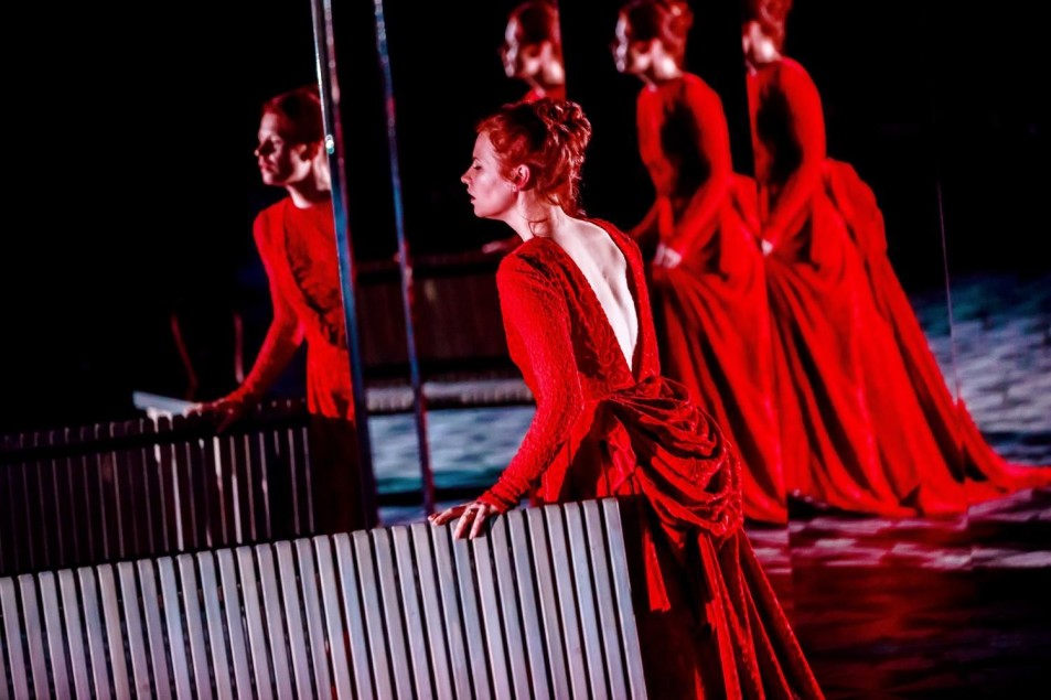Kobieta w długiej czerwonej sukni stoi przy jasnej barierce na scenie. Jej sylwetka powtarza się wielokrotnie w lustrzanych odbiciach ustawionych w tle.