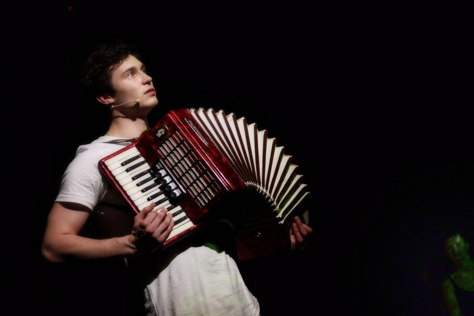 Aktor gra na czerwonym akordeonie, stojąc na ciemnej scenie. Ubrany w jasny strój, patrzy w górę. Instrument wyraźnie widoczny na tle czerni.
