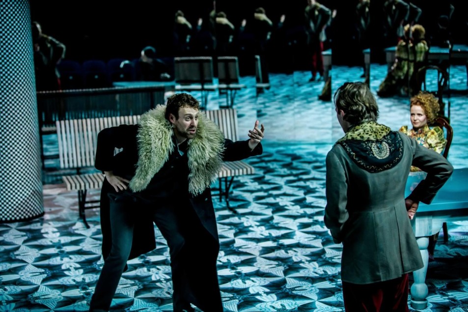 Scena teatralna: mężczyzna w ciemnym stroju z futrzastą kamizelką gestykuluje energicznie w stronę drugiego mężczyzny stojącego tyłem. W tle stylizowana przestrzeń z ławkami i lustrami, chłodna, niebiesko-zielona kolorystyka podłogi.