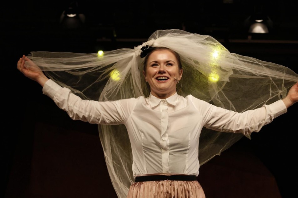 Uśmiechnięta aktorka w białej koszuli trzyma rozłożony welon nad głową. Stoi na scenie, oświetlona reflektorami.