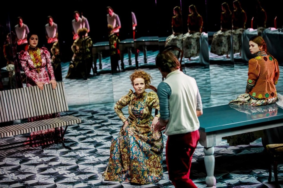 Scena zbiorowa na scenie teatralnej: kobieta w bogato zdobionej sukni siedzi przy stole, rozmawiając z mężczyzną stojącym naprzeciw. Wokół inne postacie w historycznych kostiumach, część odbita w lustrzanej ścianie, geometryczna podłoga.