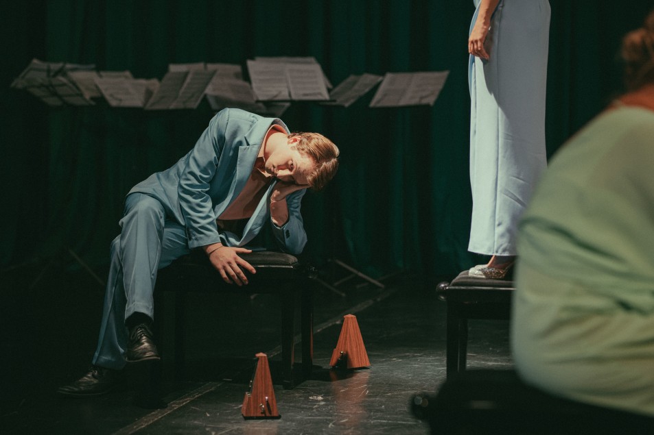 Mężczyzna w niebieskim garniturze siedzi na stołku z głową opartą na dłoni, obok stoją dwa metronomy. Z prawej strony widać nogi kobiety stojącej na innym stołku oraz fragment pulpitów z nutami w tle.