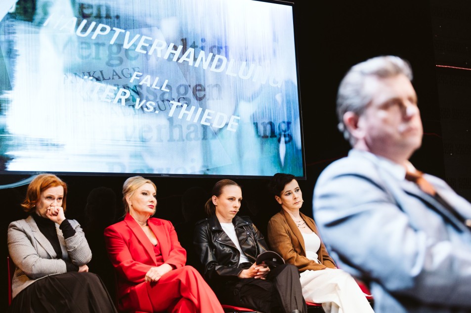 Po prawej nieostra sylwetka mężczyzny. W głębi cztery kobiety. Siedzą w rzędzie na czerwonych krzesłach. Nad nimi ekran z projekcją słów po niemiecku.