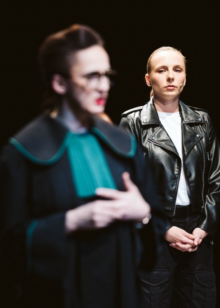 Przodem do nas brunetka w todze adwokackiej (Anna Kociarz). W głębi młoda kobieta w czarnej ramonesce (Karolina Winarska). Patrzy na nią. 