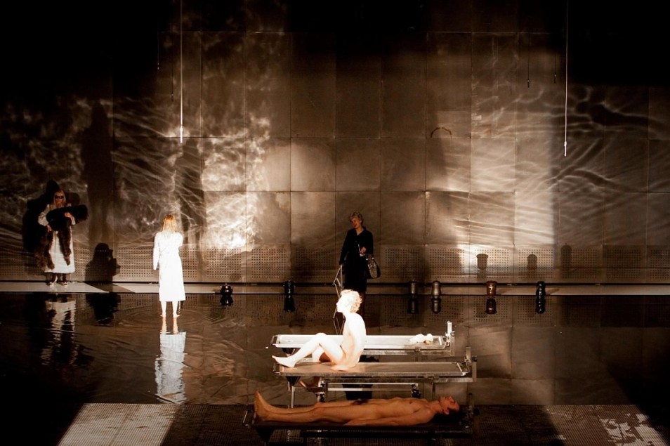 Na zalanej wodą scenie jedna osoba siedzi naga na metalowym stole, druga leży pod nim. W tle stoją 3 kobiety, obserwujące scenę w teatralnym świetle.