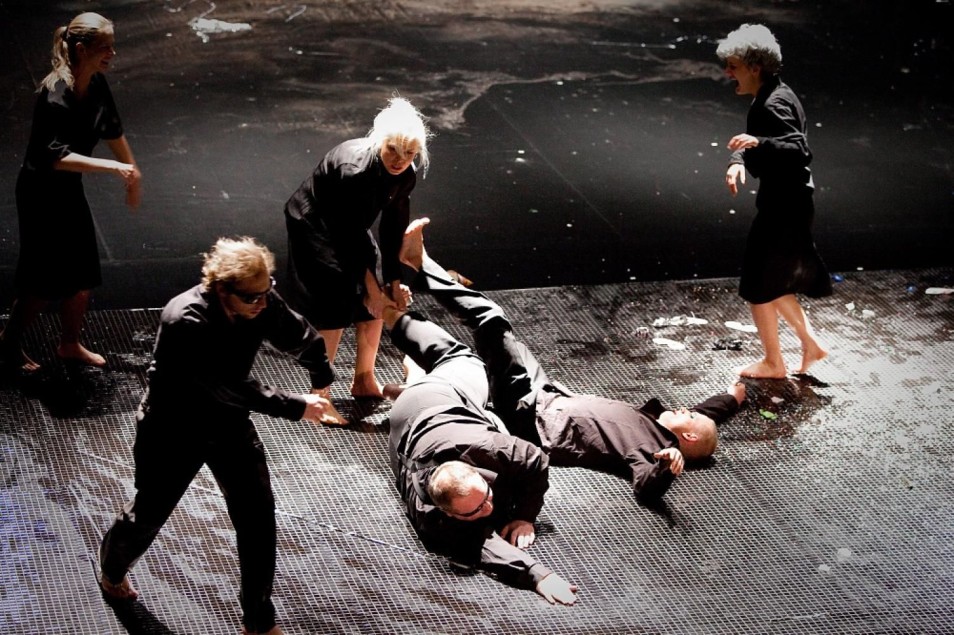 Kilka osób w czarnych kostiumach porusza się po mokrej scenie. Dwie postacie leżą na podłodze, inne pochylają się nad nimi w dynamicznej, chaotycznej akcji.