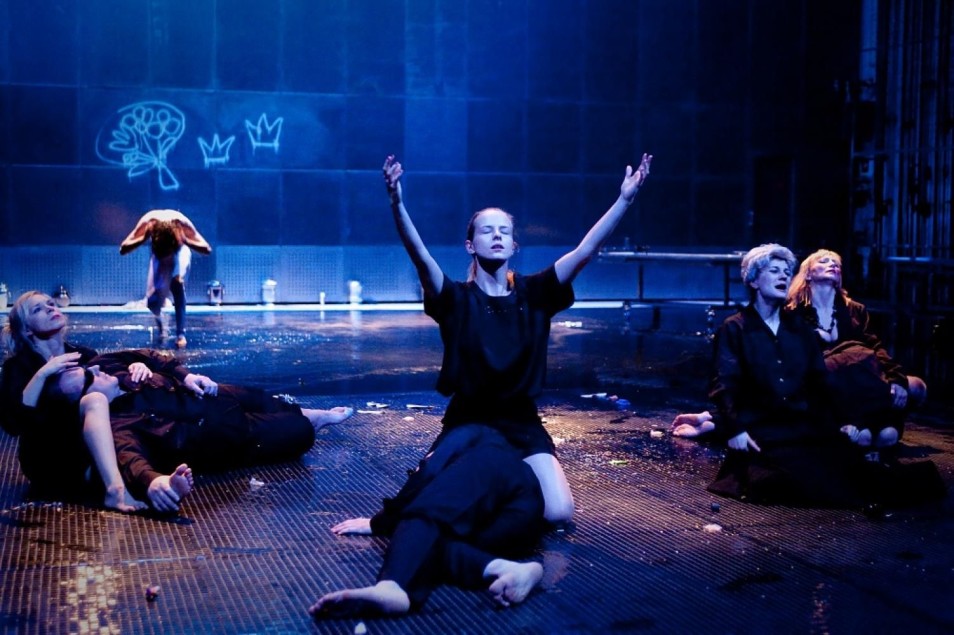 Na niebiesko oświetlonej scenie grupa performerów klęczy i siedzi na podłodze. Centralna postać unosi ręce ku górze, pozostali leżą lub opierają się o siebie.