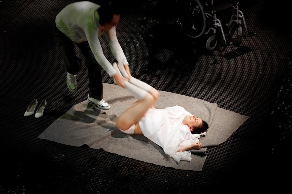 Mężczyzna pochyla się nad kobietą leżącą na macie i unosi jej nogi. Kobieta ubrana jest na biało, obok stoją buty i wózek inwalidzki na ciemnej scenie.
