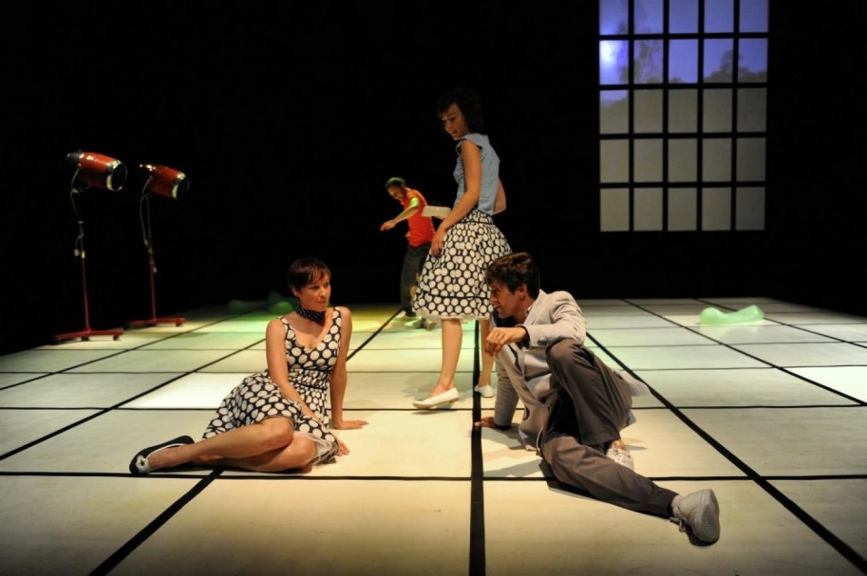 Kilka osób na scenie w różnych pozach siedzących i stojących. Minimalistyczna scenografia, skupienie na relacjach między postaciami.