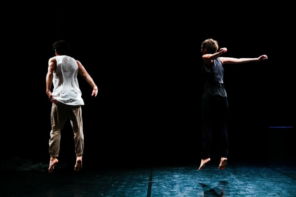 Dwójka tancerzy skacze jednocześnie na scenie, widok od tyłu. Oboje boso, ramiona rozłożone, uchwycony moment w powietrzu na tle ciemnej sceny.