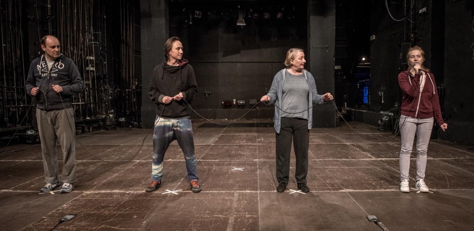 Czworo aktorów stoi na scenie w jednej linii, trzymając przewody w dłoniach. Przestrzeń teatralna bez scenografii, atmosfera próby lub performansu.
