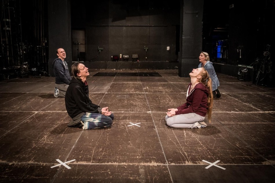 Czworo aktorów klęczy na podłodze sceny, z uniesionymi twarzami i dłońmi. Układ symetryczny, gesty sugerują emocjonalne napięcie lub modlitwę.