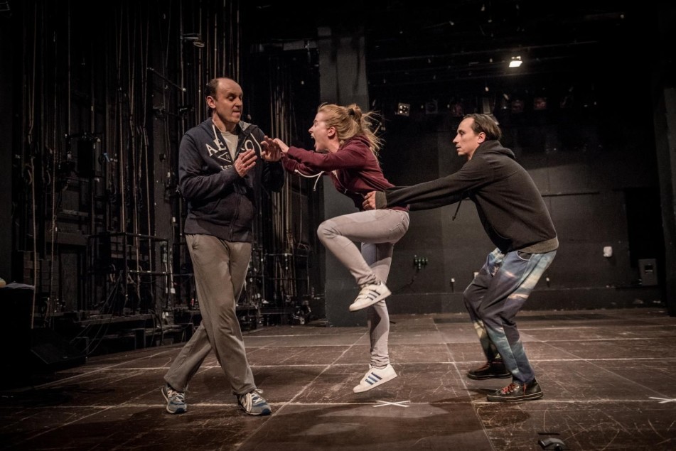 Troje aktorów w dynamicznej scenie: kobieta wyskakuje do przodu, krzycząc, powstrzymywana przez mężczyznę. Drugi mężczyzna reaguje gestem obronnym.