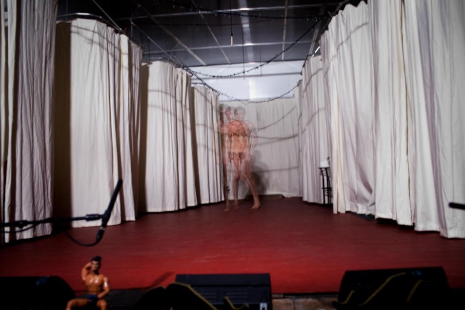 Scena z białymi zasłonami po bokach. Na czerwonej podłodze widać rozmytą sylwetkę nagiego performera, uchwyconą w ruchu, co tworzy efekt wielokrotnego obrazu.